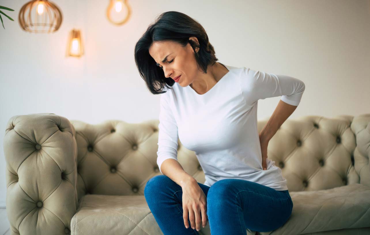 woman holding lower back while sitting on edge of couch