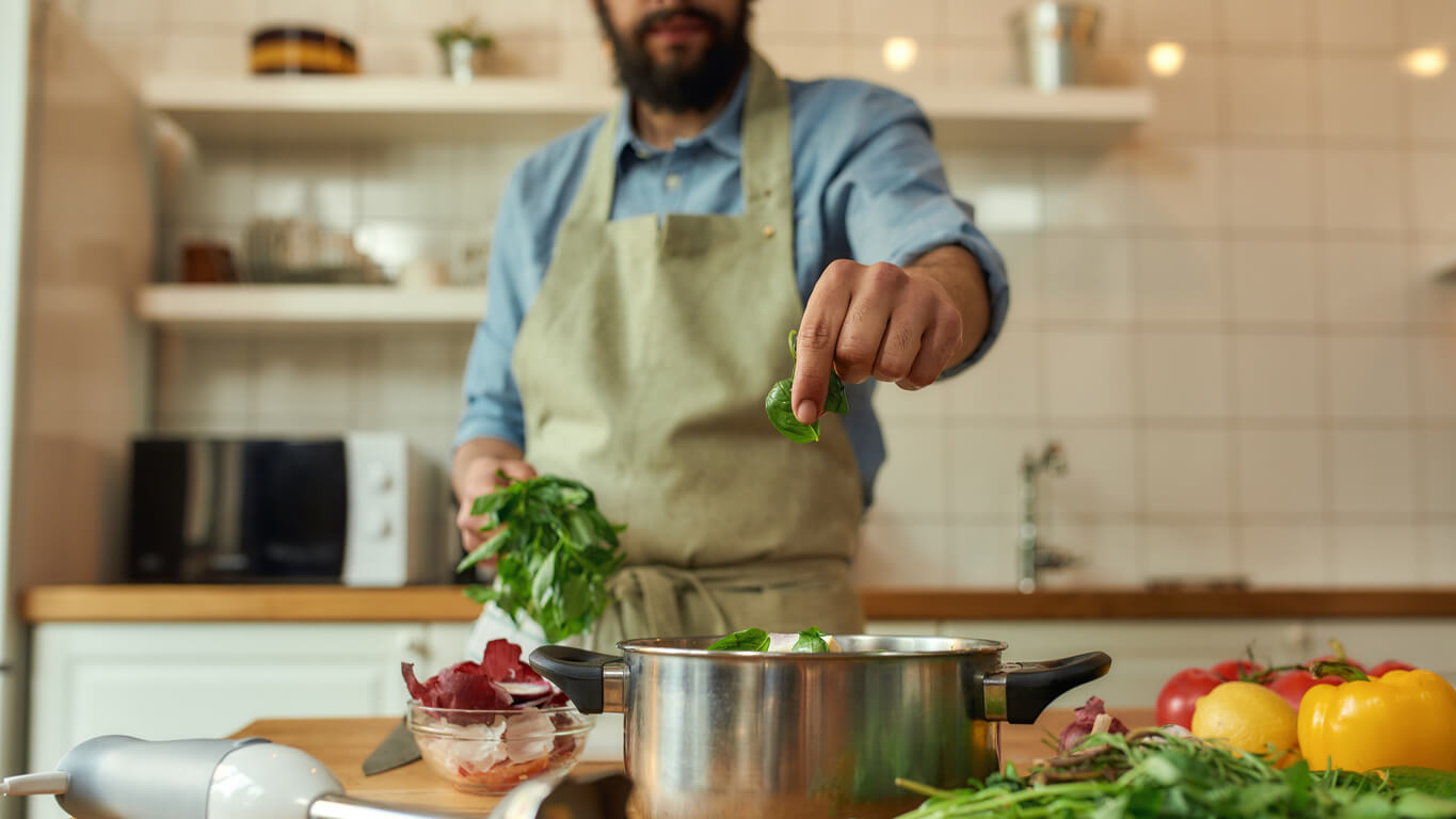cropped shot of man chef cook adding basil leaf to the pot with chopped vegetables